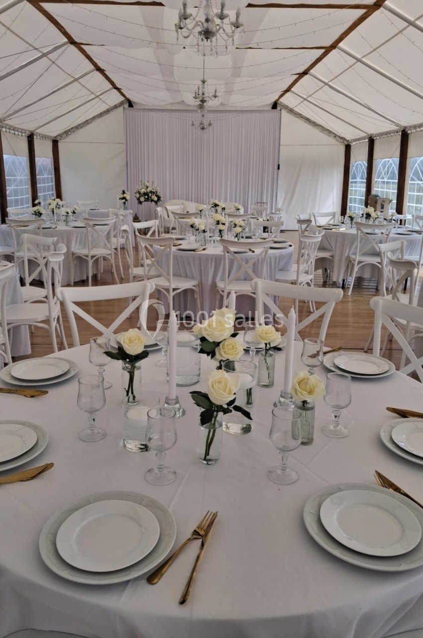 Salle de réception décorée avec des tables rondes dressées, nappes blanches, roses blanches et couverts dorés.