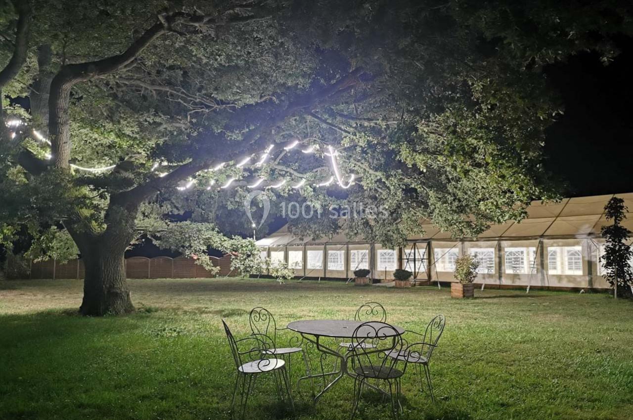 Un grand arbre illuminé la nuit près d'une tente éclairée, avec une table et des chaises en métal sur la pelouse.