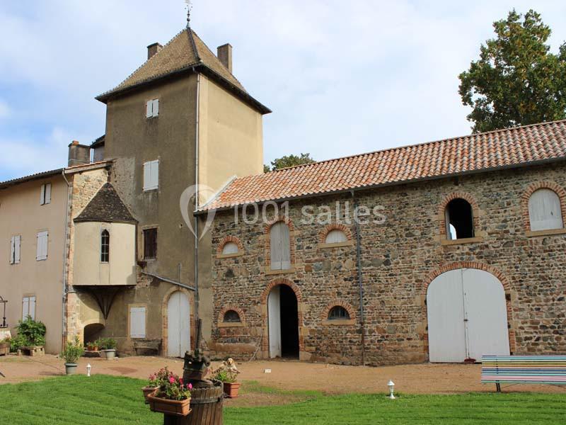 Bâtiment en pierre et crépi avec une tour, des volets fermés, une cour aménagée et des jardinières fleuries.