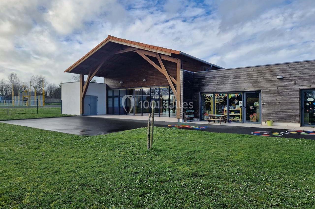 Bâtiment moderne en bois avec une entrée vitrée, entouré d'une pelouse et d'une aire de jeux visible à gauche.