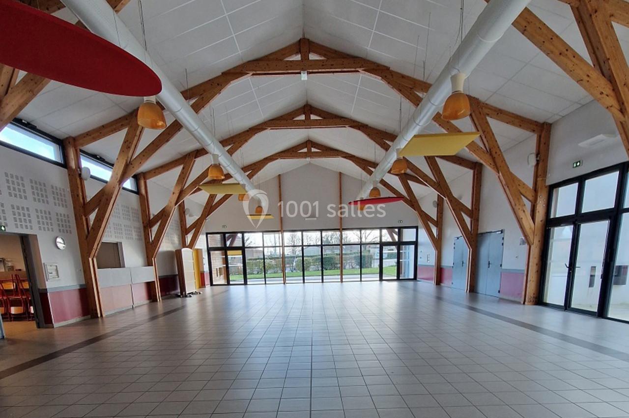 Salle polyvalente lumineuse avec charpente en bois apparente, grandes baies vitrées et carrelage au sol.