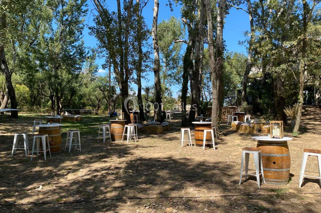 Tables en tonneaux et tabourets blancs disposés en plein air sous des arbres dans un cadre naturel ensoleillé.