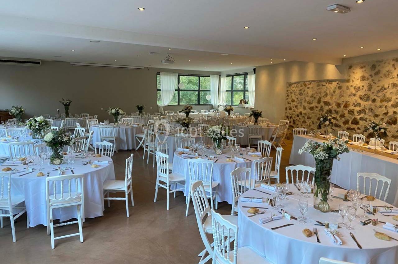 Salle de réception décorée avec des tables rondes, nappes blanches, chaises en bois et arrangements floraux.