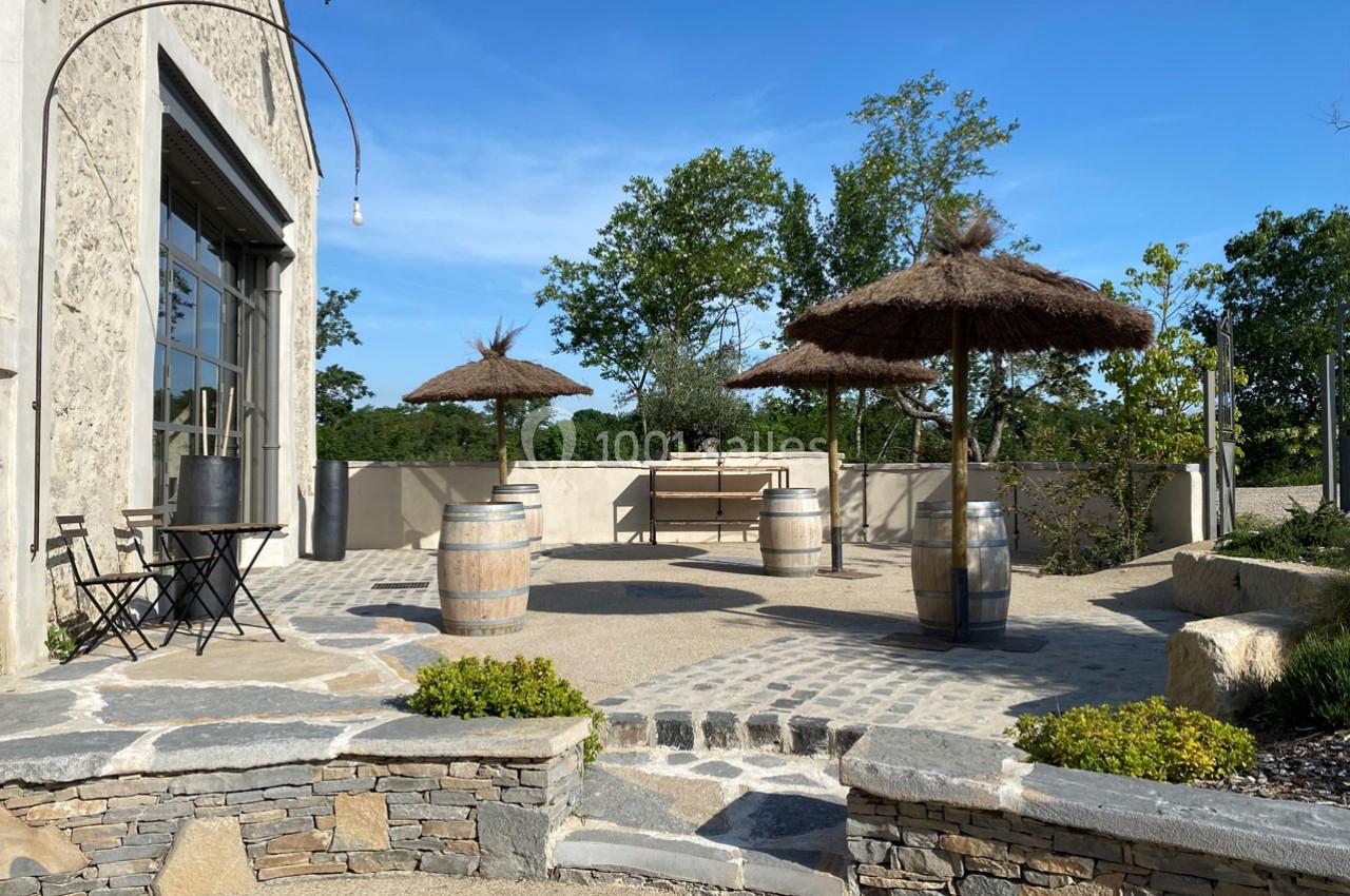 Terrasse ensoleillée avec tables, chaises, tonneaux en bois et parasols en paille, entourée de verdure.