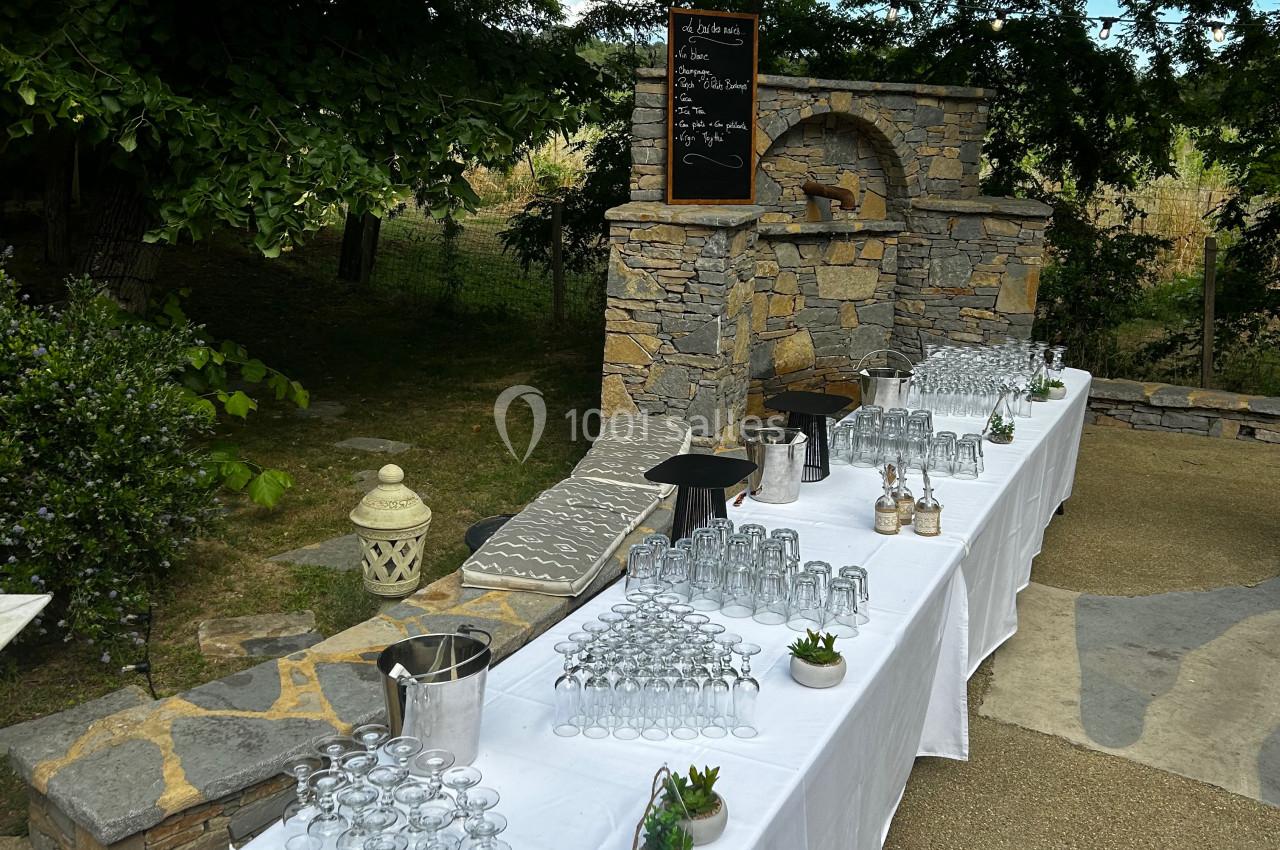 Tables dressées en extérieur avec des verres, carafes et décorations, devant un mur en pierre et un tableau noir.