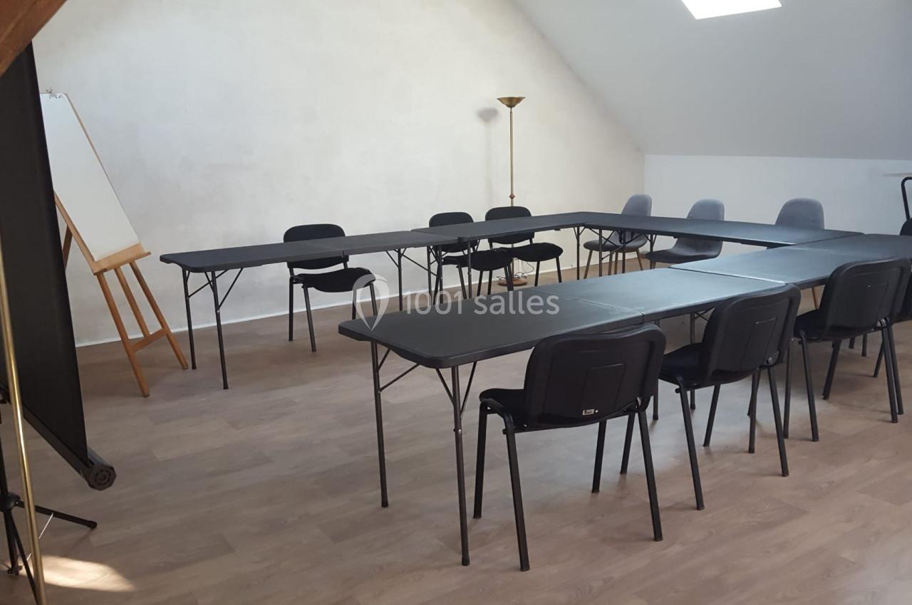 Salle de réunion lumineuse avec tables noires disposées en U, chaises noires et grises, tableau chevalet et lampadaire.