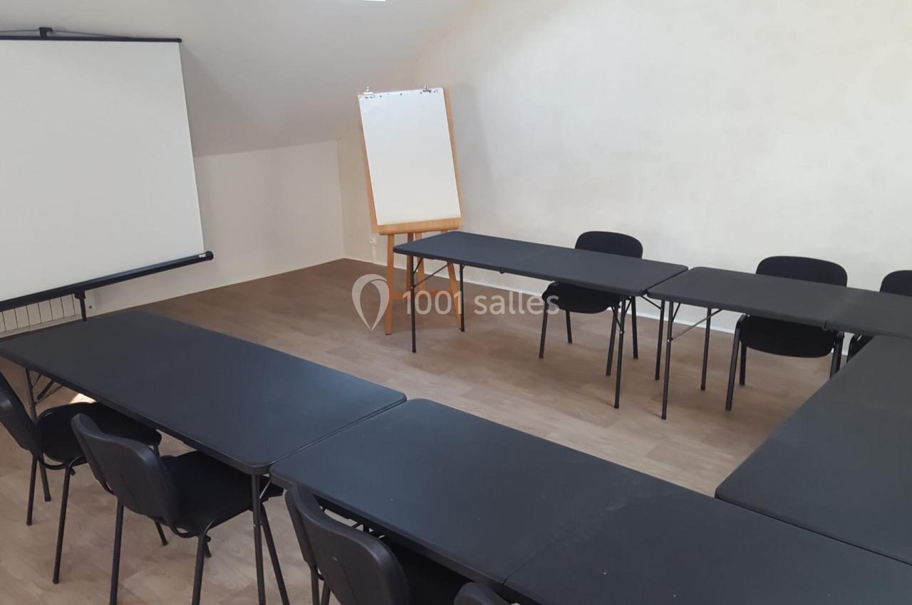 Salle de réunion avec tables noires disposées en U, chaises noires, écran de projection et tableau blanc sur chevalet.
