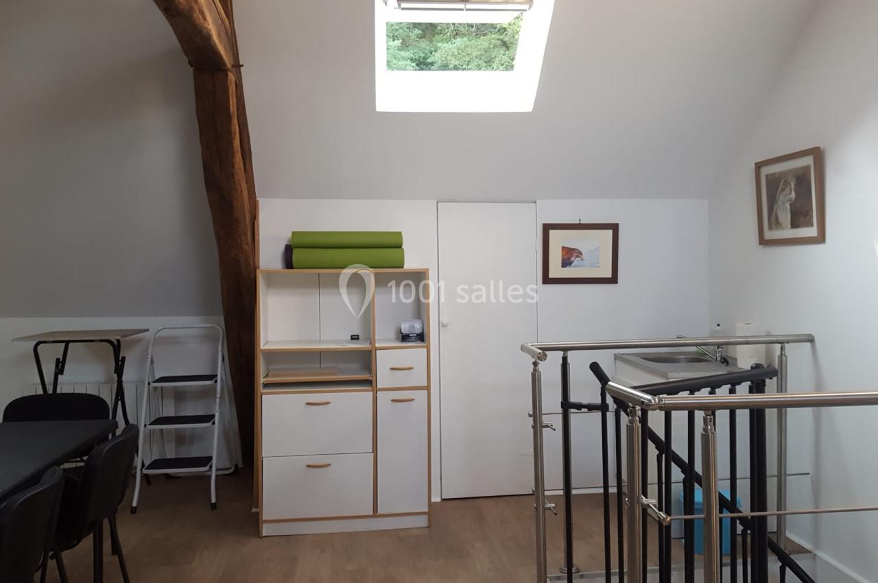 Salle lumineuse sous les combles avec poutre apparente, meubles de rangement, table, chaises et rampe d'escalier.