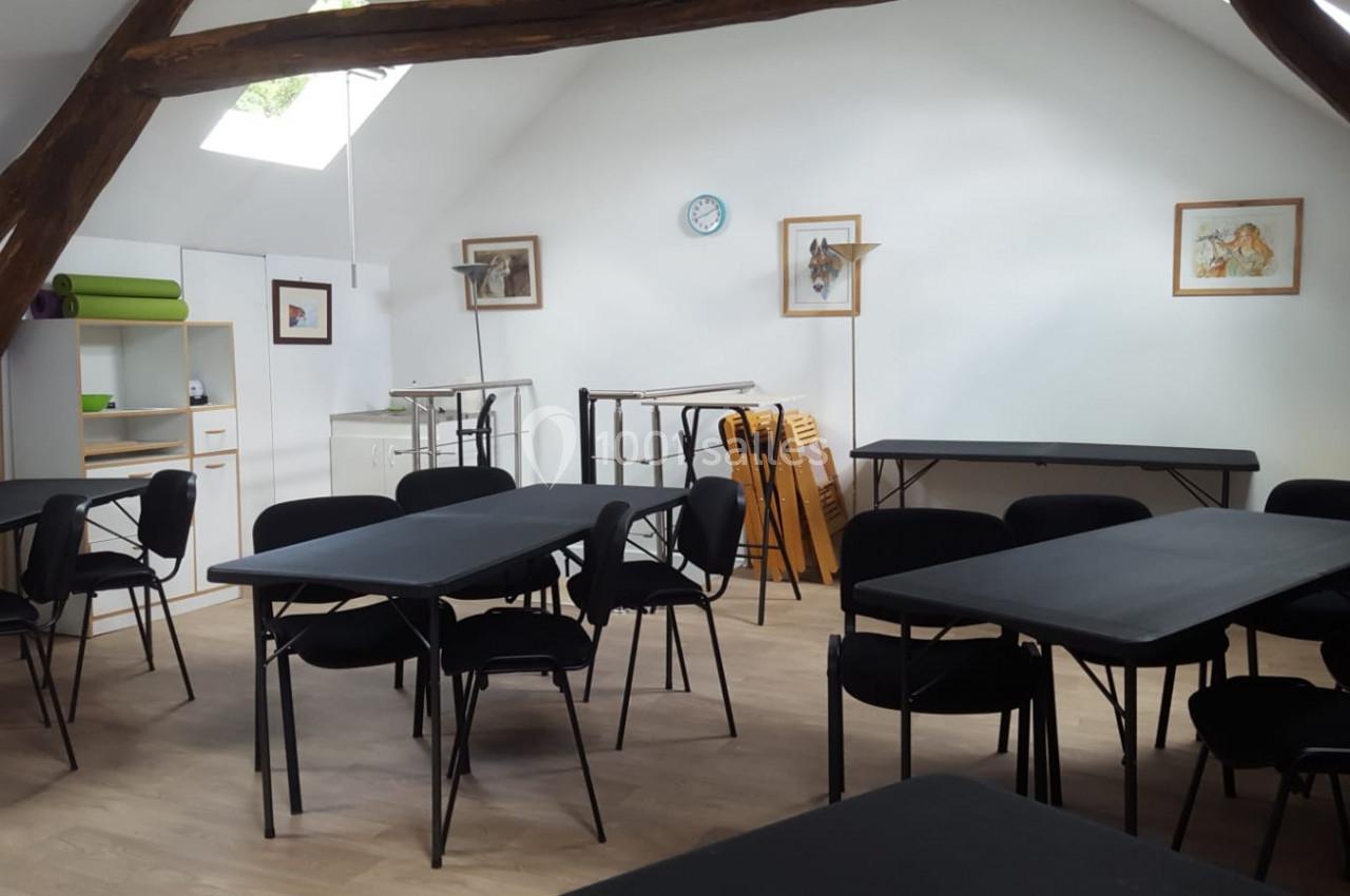 Salle lumineuse avec poutres apparentes, tables noires, chaises noires et mobilier de rangement en arrière-plan.