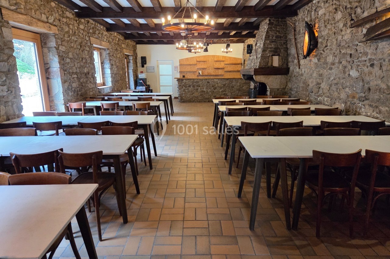 Salle à manger rustique avec murs en pierre, tables alignées et plafond en poutres apparentes.