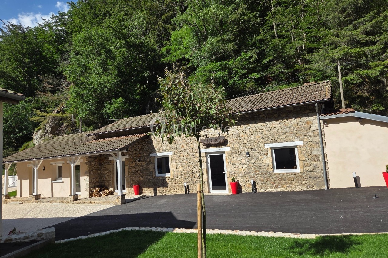 Façade d'une maison en pierre avec toit en tuiles, entourée de verdure et d'une allée asphaltée.