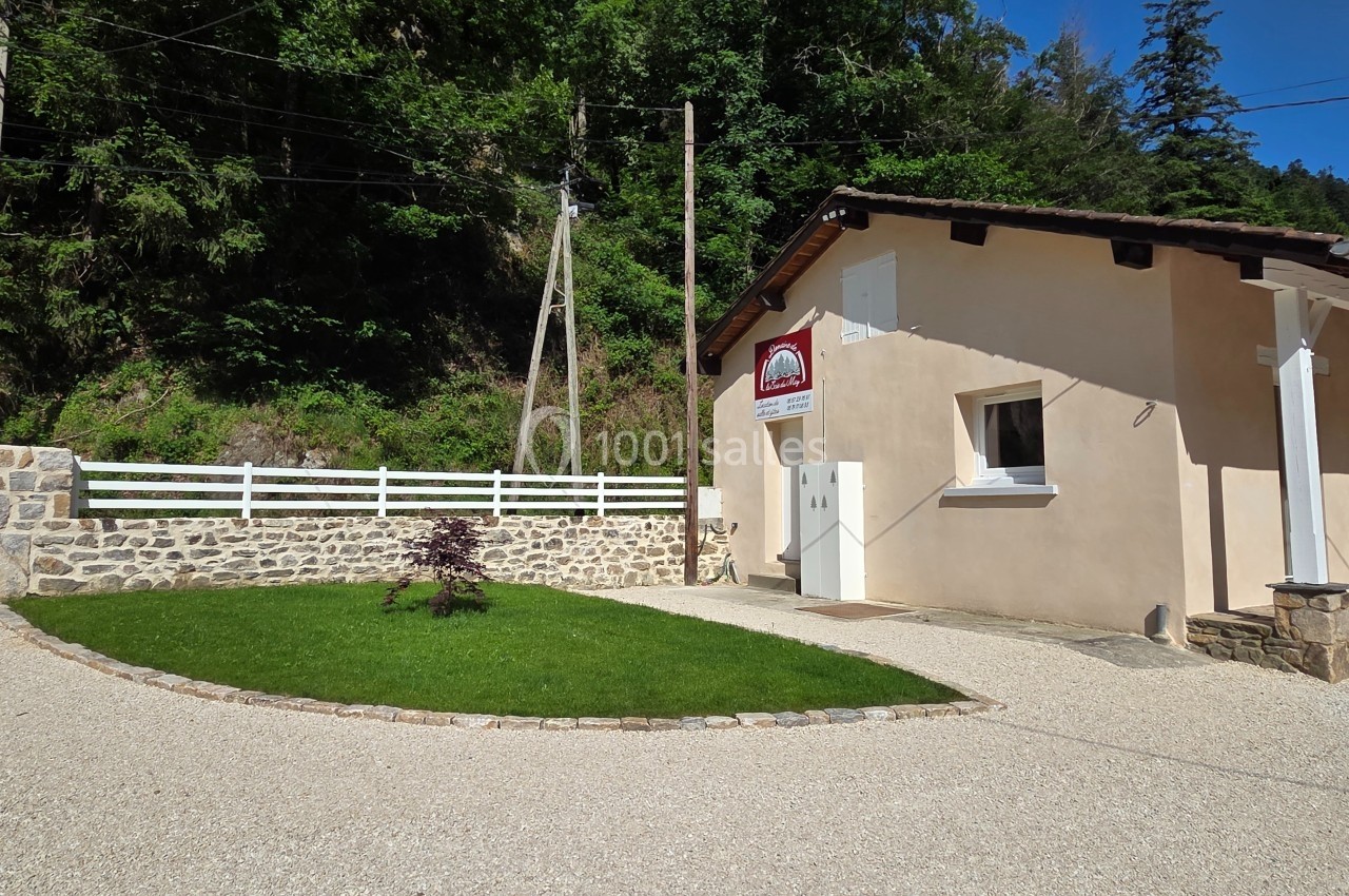 Bâtiment clair avec enseigne, entouré d'une pelouse, d'un chemin gravillonné et d'une clôture en bois devant une forêt.