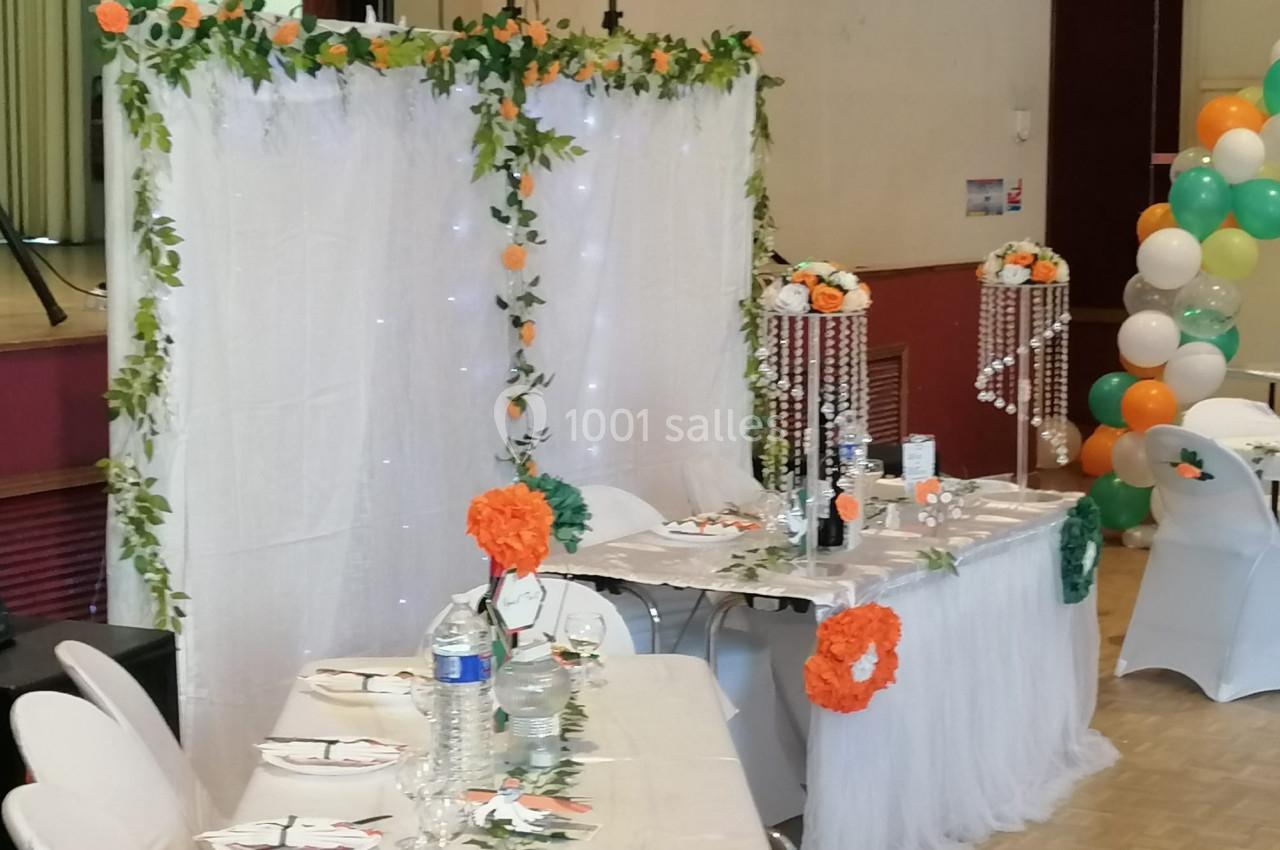 Décoration de table de fête avec nappes blanches, fleurs orange, guirlandes lumineuses et ballons colorés en arrière-plan.
