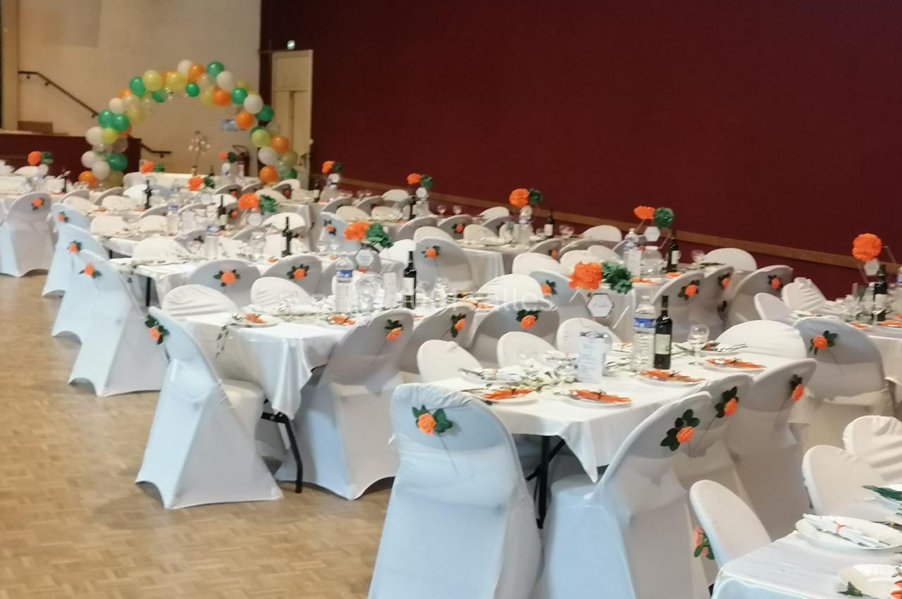 Salle de réception décorée avec des tables dressées, nappes blanches, chaises assorties et ballons colorés en arrière-plan.