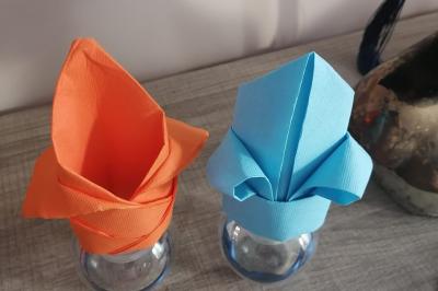 Serviettes pliées en forme de fleurs, orange et bleue, disposées sur des verres transparents sur une table en bois.