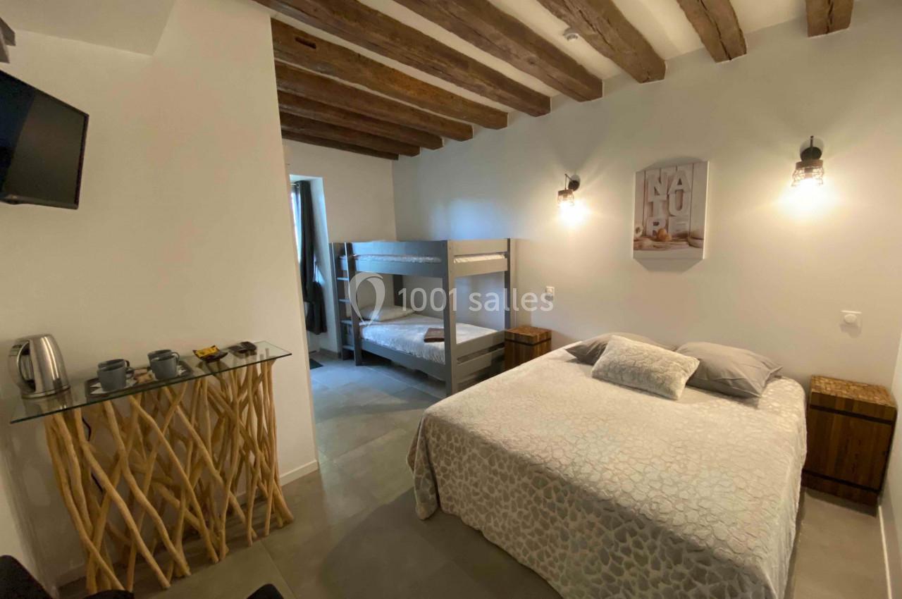 Chambre avec lit double, lits superposés, table en bois, télévision murale et éclairage mural sous plafond à poutres…