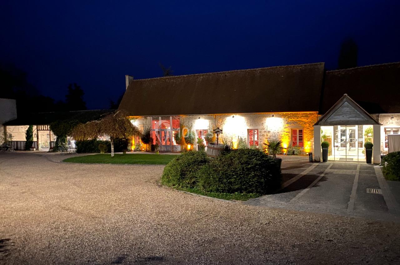 Bâtiment rural éclairé de nuit avec des murs en pierre, des fenêtres rouges et une entrée vitrée sur la droite.