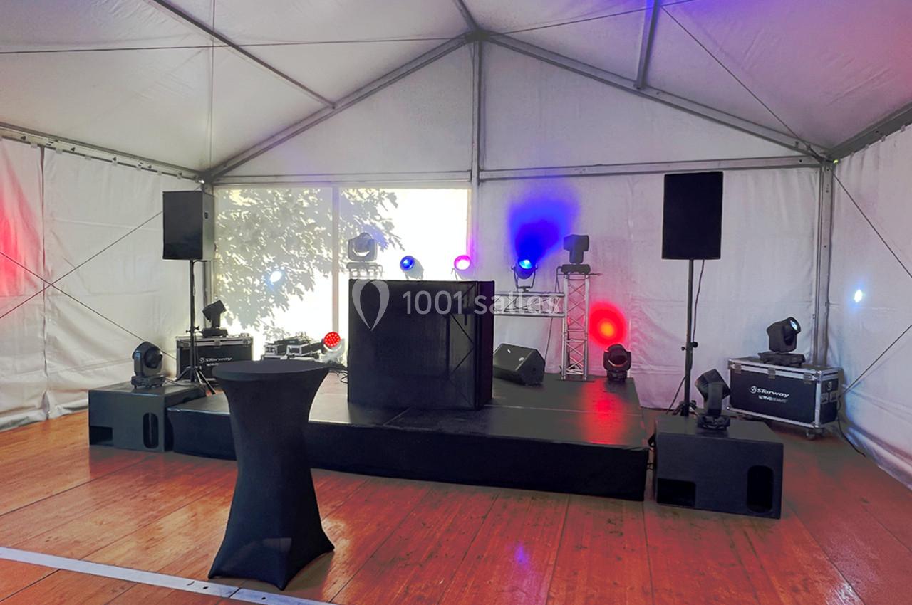 Scène avec équipement de sonorisation et éclairage coloré dans une tente blanche sur un sol en bois.
