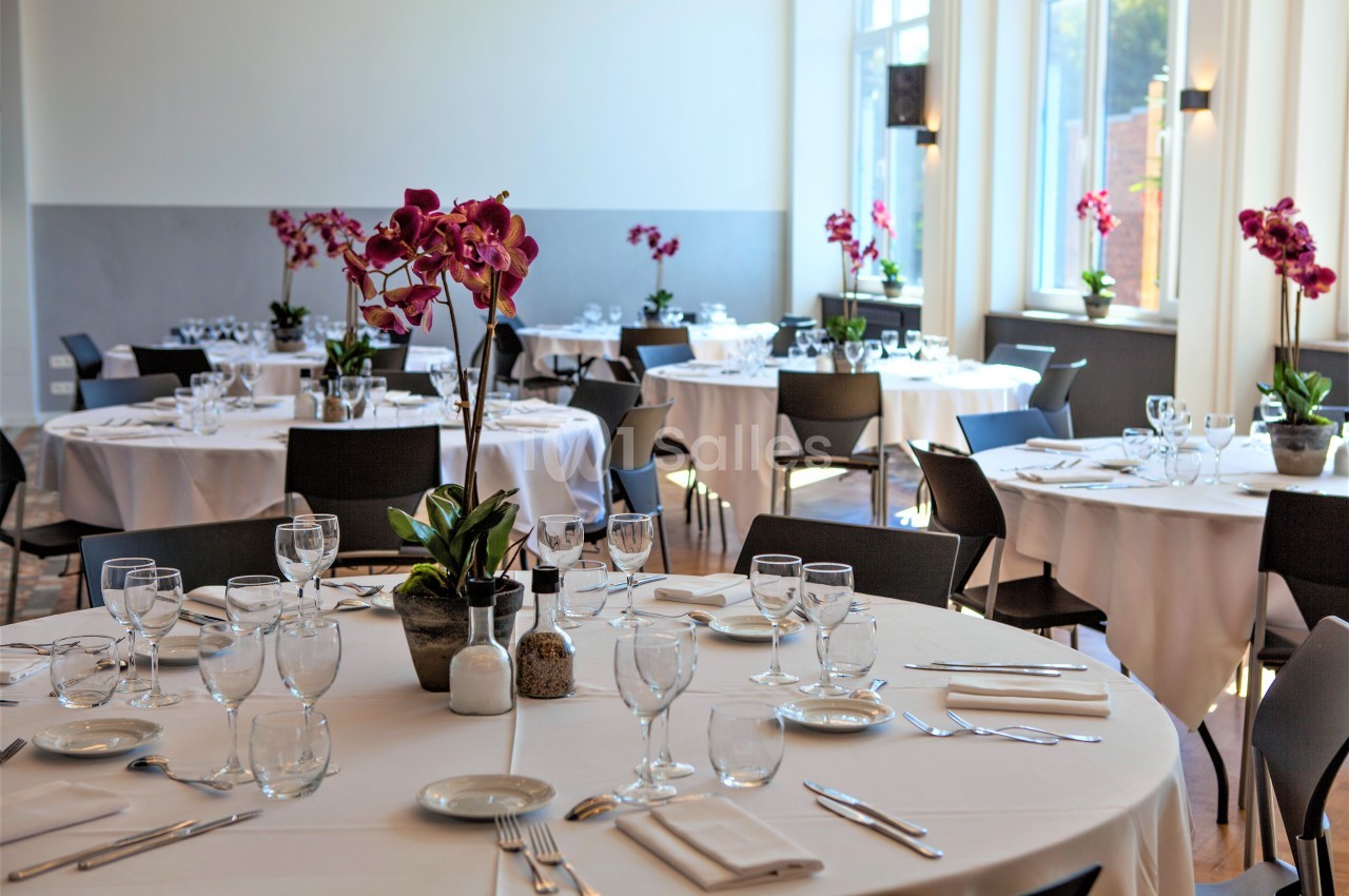 Salle de réception lumineuse avec tables rondes dressées, nappes blanches et orchidées en centre de table.