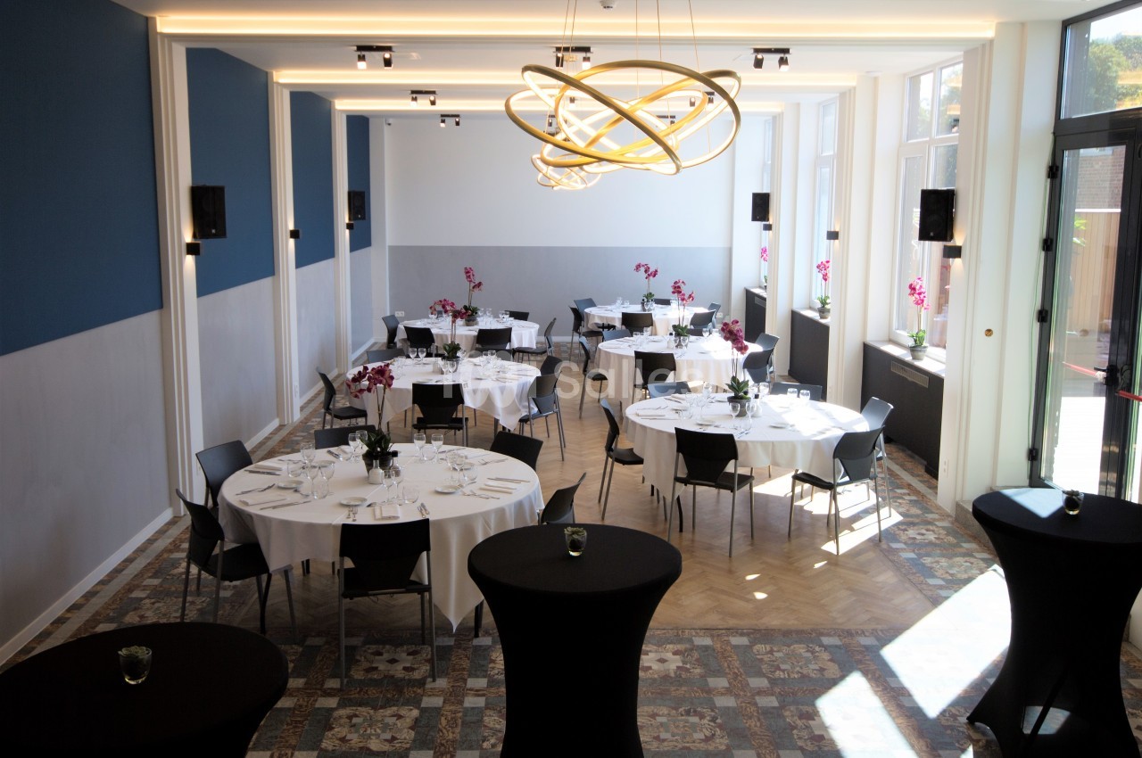 Salle lumineuse aménagée pour un événement, avec tables rondes dressées, chaises noires et décorations florales.