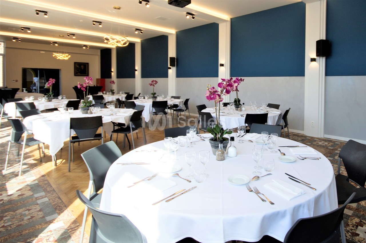 Salle de réception lumineuse avec tables rondes dressées, nappes blanches et orchidées en centre de table.