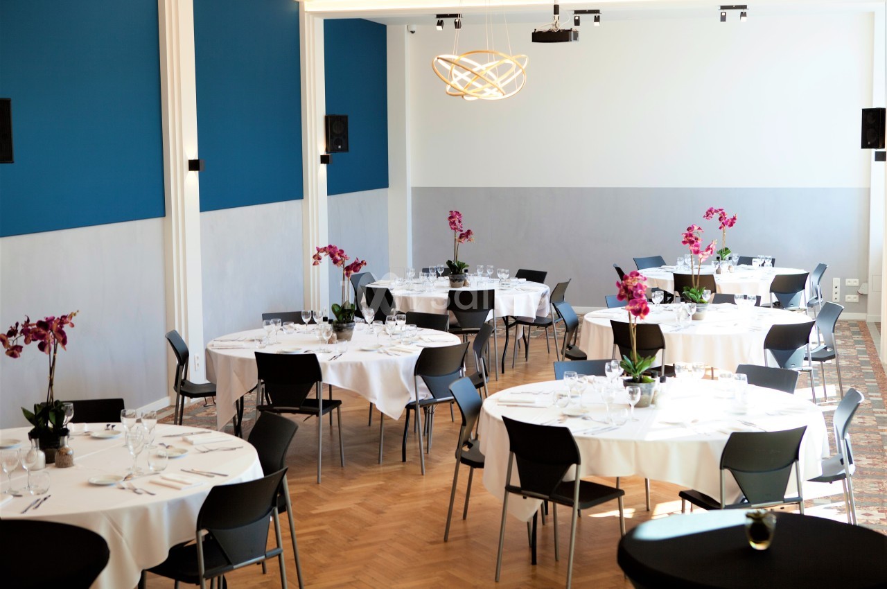 Salle de réception lumineuse avec tables rondes dressées, nappes blanches et décorations florales violettes.
