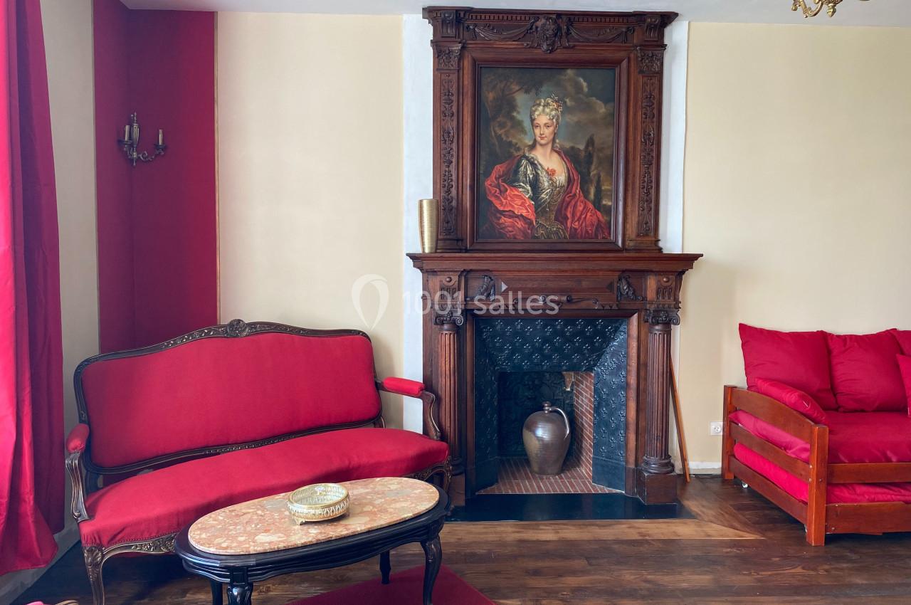 Salon avec mobilier ancien, canapé et fauteuil rouges, cheminée en bois sculpté surmontée d'un portrait.