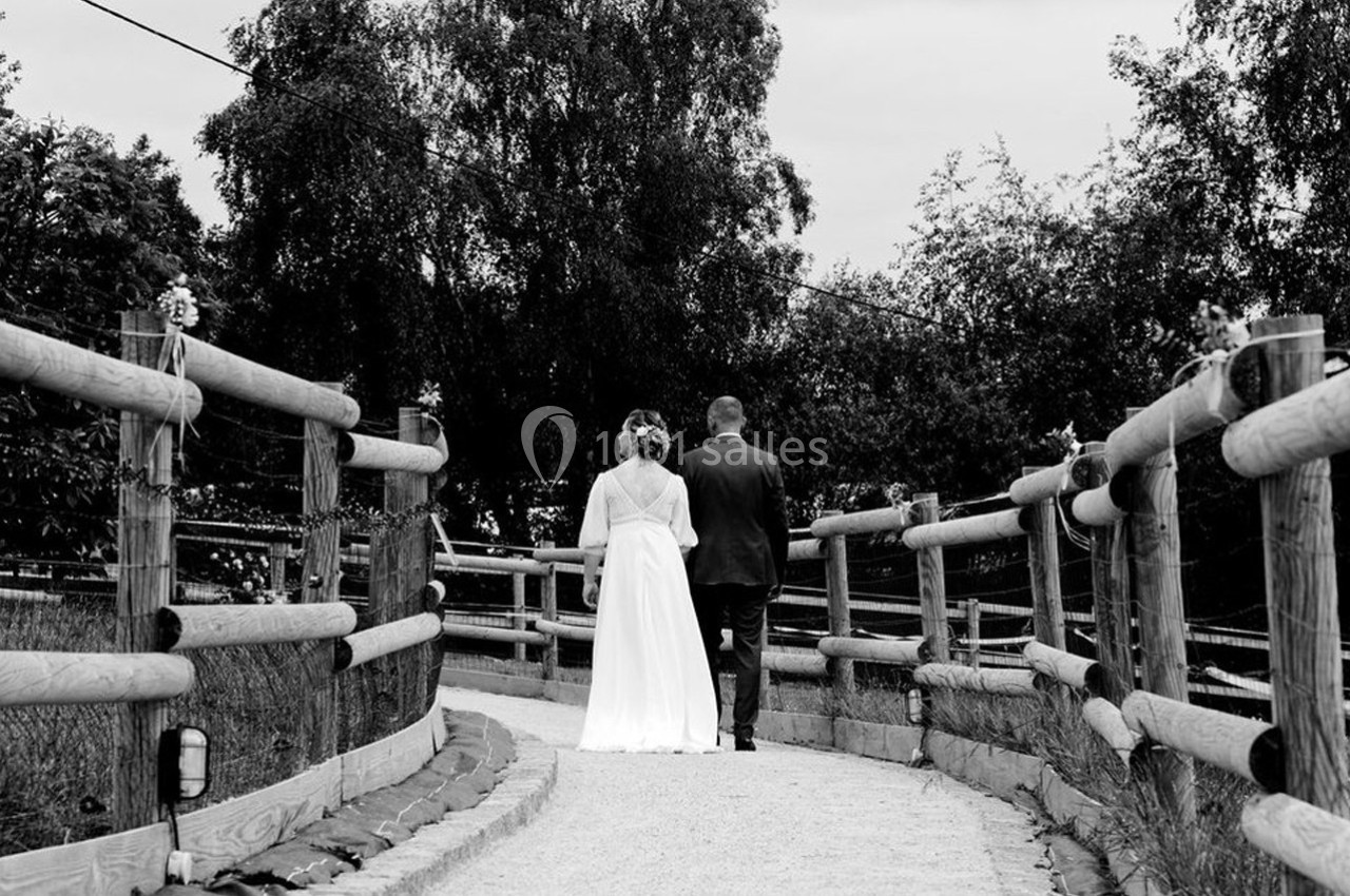 Un couple en tenue de mariage marche sur un chemin bordé de barrières en bois, entouré de végétation.