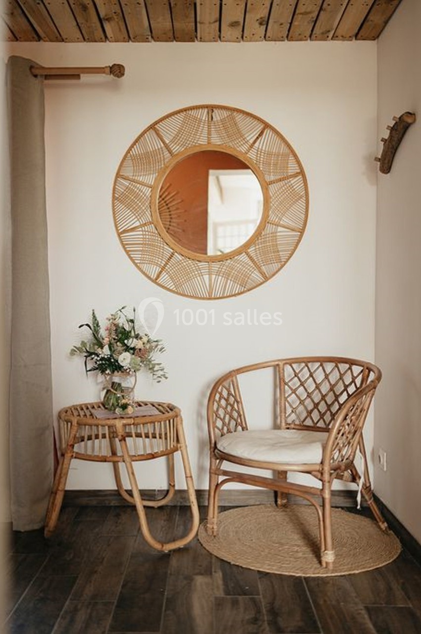 Coin décoratif avec fauteuil en rotin, table assortie, miroir rond tressé et bouquet de fleurs, sur sol en bois.