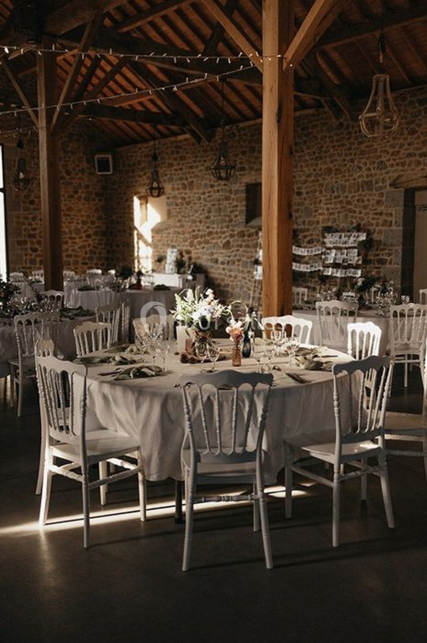 Salle de réception en pierre avec tables rondes dressées, nappes blanches et chaises blanches, éclairée par une lumière…