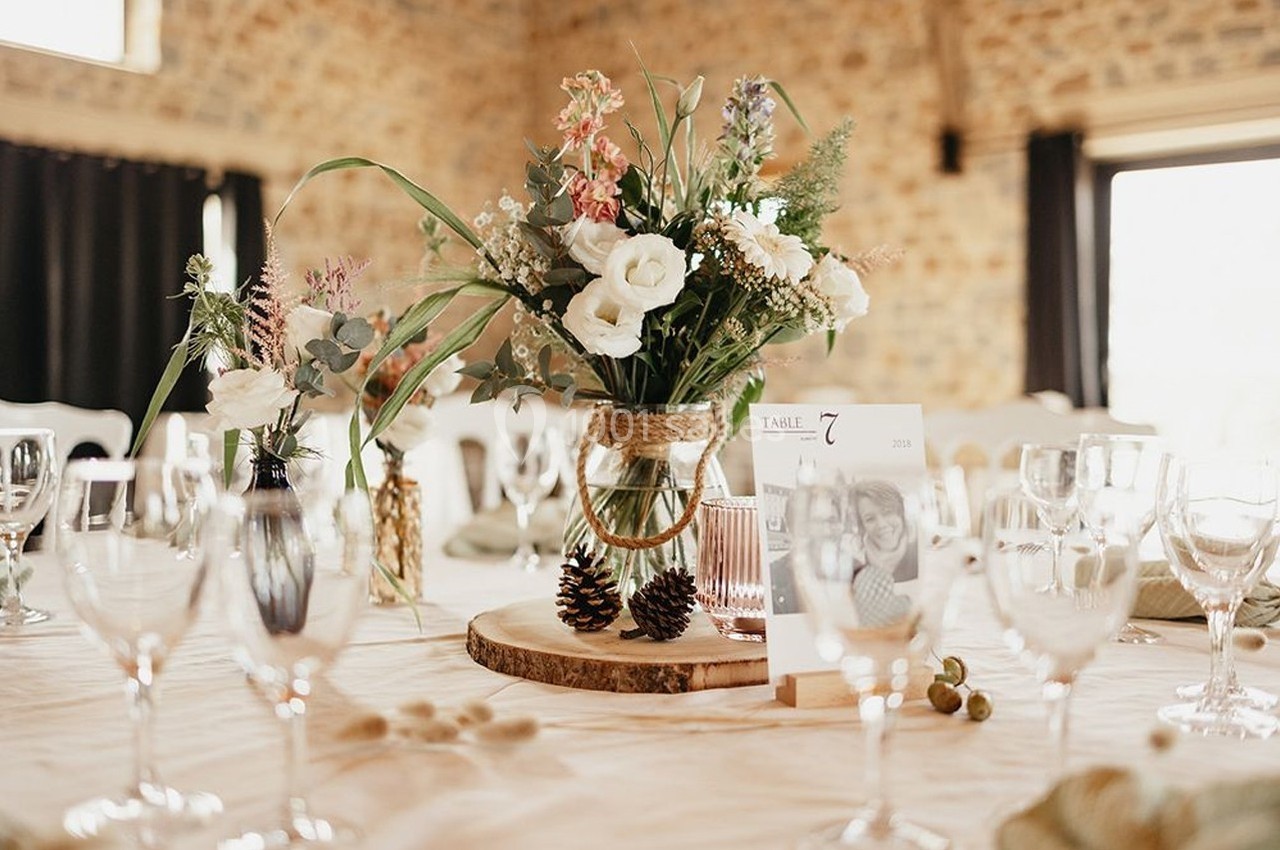 Centre de table floral avec des fleurs blanches et colorées, posé sur une rondelle de bois, entouré de verres et de…