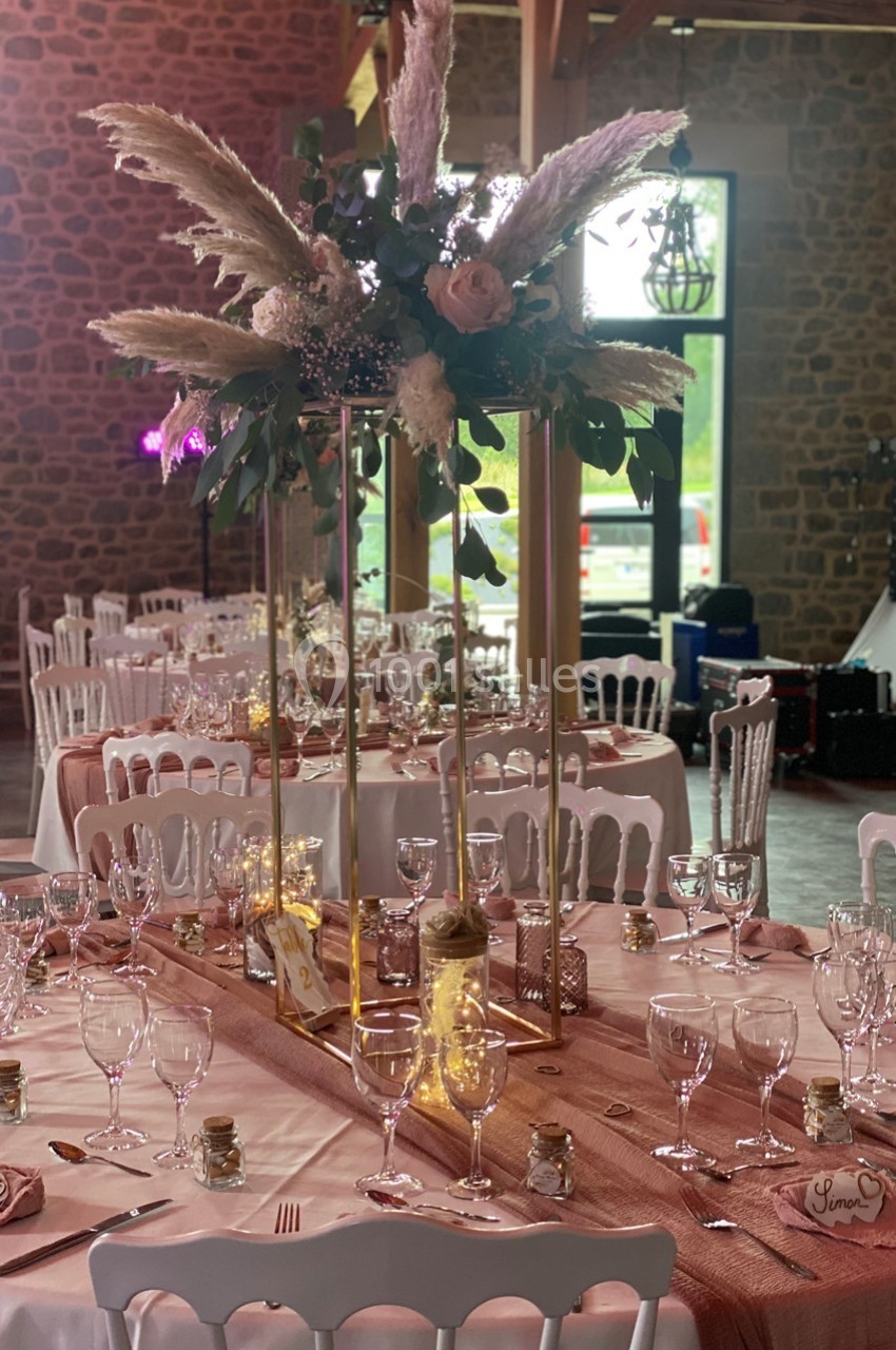 Salle de réception décorée avec des tables dressées, centre de table floral et nappes roses dans un cadre rustique.