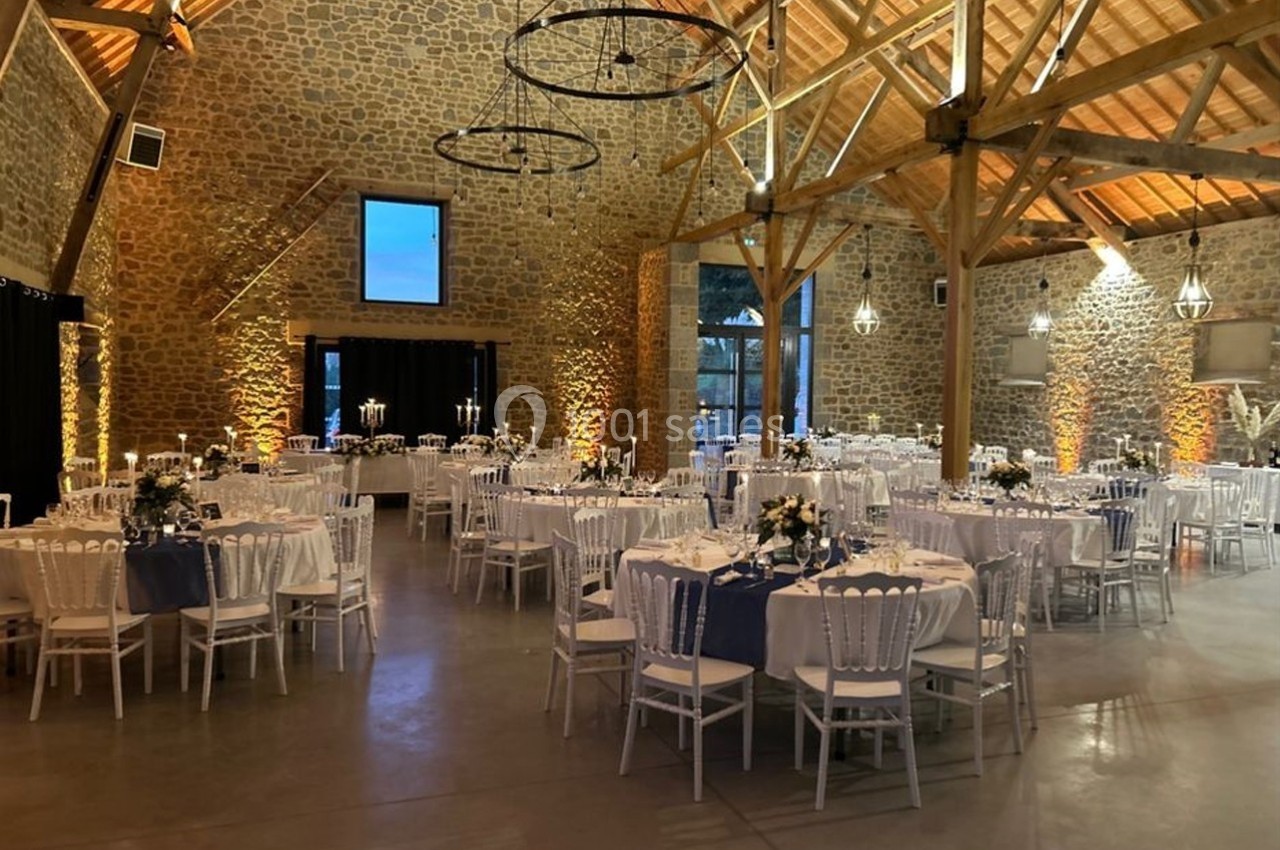 Salle de réception en pierre avec tables rondes dressées, chaises blanches et éclairage chaleureux.