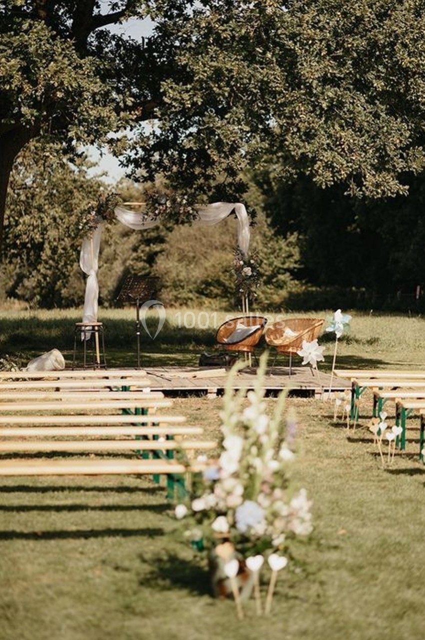 Chaises et arche décorée pour une cérémonie en plein air, entourées de bancs en bois et de verdure.