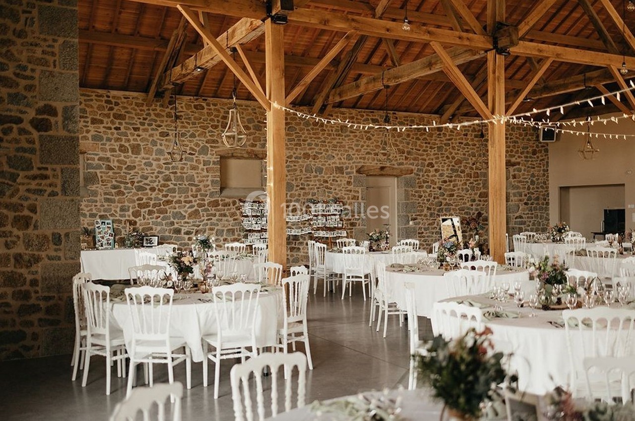 Salle de réception avec murs en pierre, poutres apparentes, tables rondes décorées et chaises blanches.