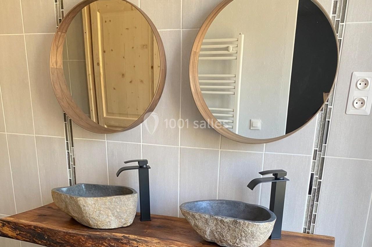 Deux vasques en pierre avec robinets noirs sur un plan en bois brut, devant deux miroirs ronds.