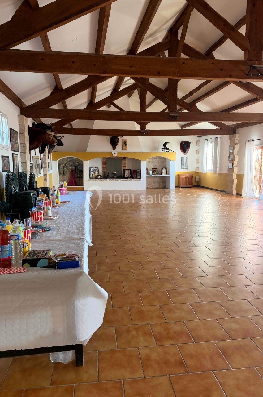 Grande salle avec charpente en bois apparente, tables dressées sur la gauche et sol en carrelage orange.
