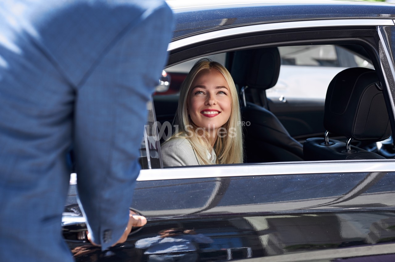 Une femme souriante assise dans une voiture noire regarde un homme en costume debout à l'extérieur.