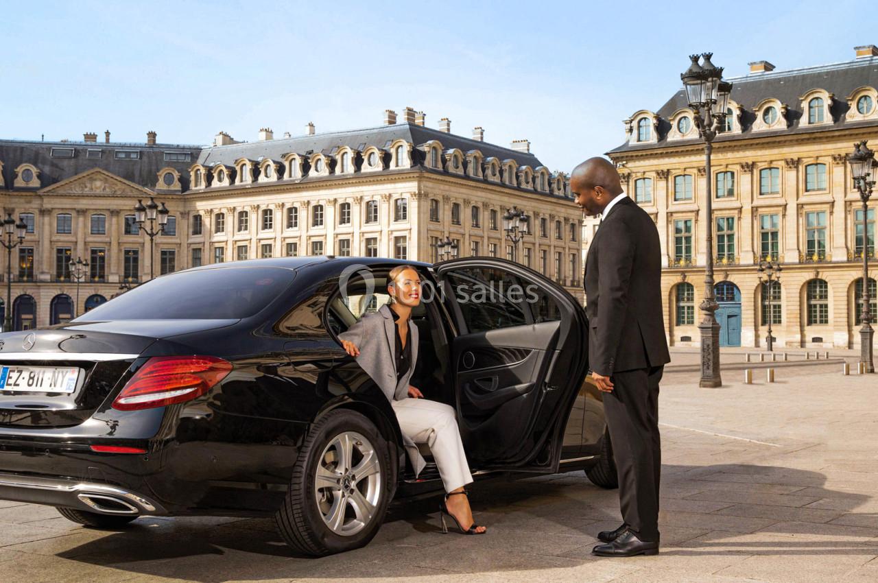 Une femme sort d'une voiture noire avec un chauffeur en costume, sur une place bordée de bâtiments élégants.