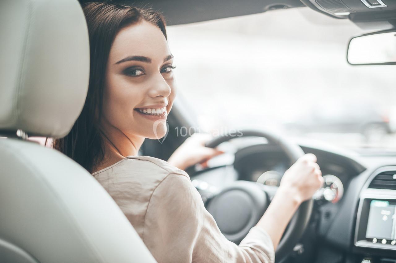 Une femme souriante conduit une voiture, vue depuis le siège passager.