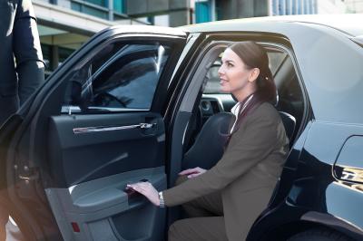 Une femme en tenue professionnelle sort d'une voiture noire, accueillie par un chauffeur en costume.