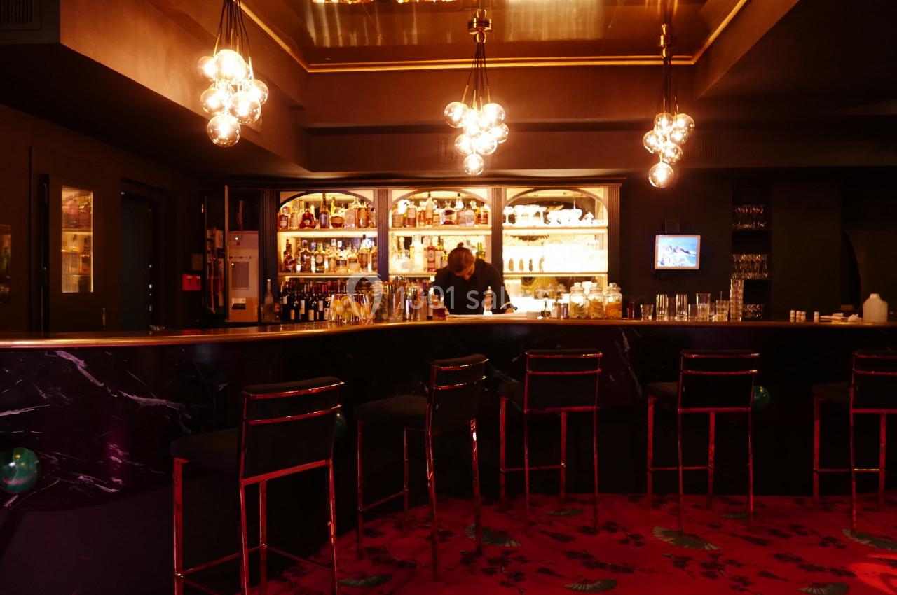 Bar élégant avec comptoir en marbre noir, étagères éclairées remplies de bouteilles et tabourets alignés sur un tapis rouge.