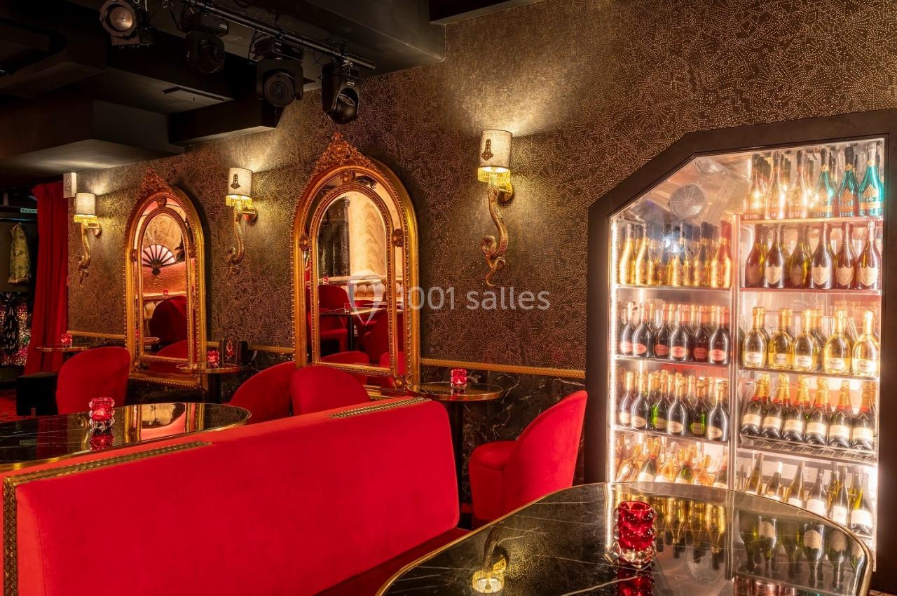Salle élégante avec fauteuils rouges, tables en marbre, miroirs dorés et réfrigérateur rempli de bouteilles.