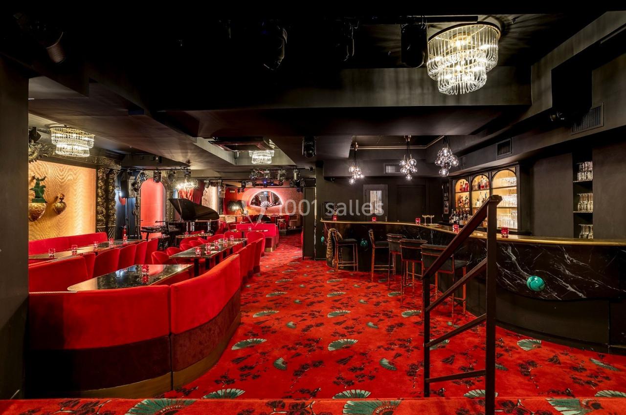 Salle de spectacle élégante avec fauteuils rouges, tapis rouge à motifs, bar en marbre noir et lustres suspendus.