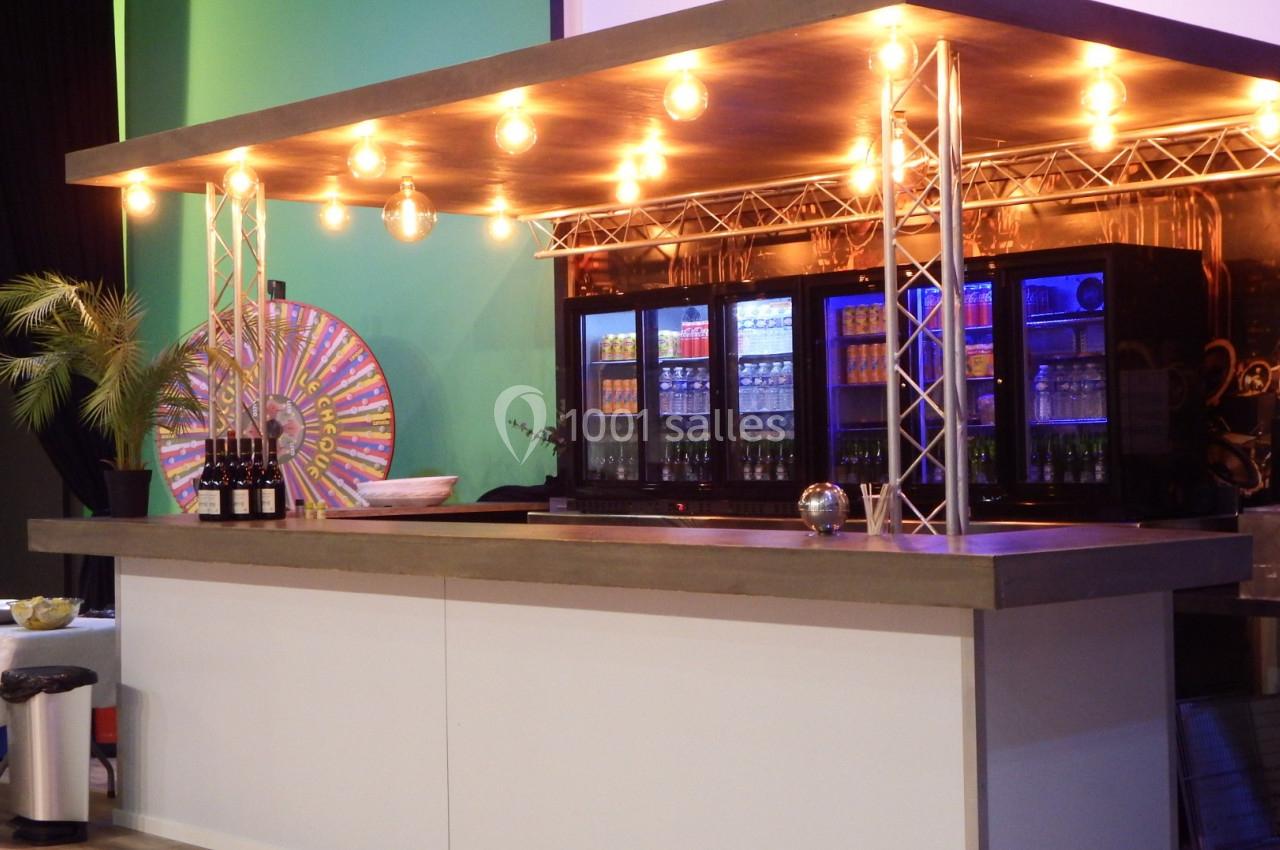 Comptoir de bar moderne avec éclairage suspendu, réfrigérateurs remplis de boissons et une roue colorée en arrière-plan.