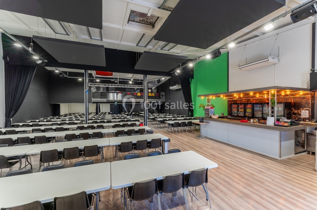 Salle spacieuse avec tables et chaises alignées, comptoir éclairé à droite et fond vert sur un mur au fond.