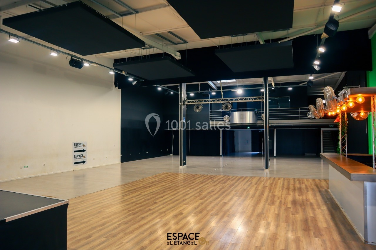 Salle polyvalente avec parquet, espace scénique, mezzanine métallique et éclairage suspendu.