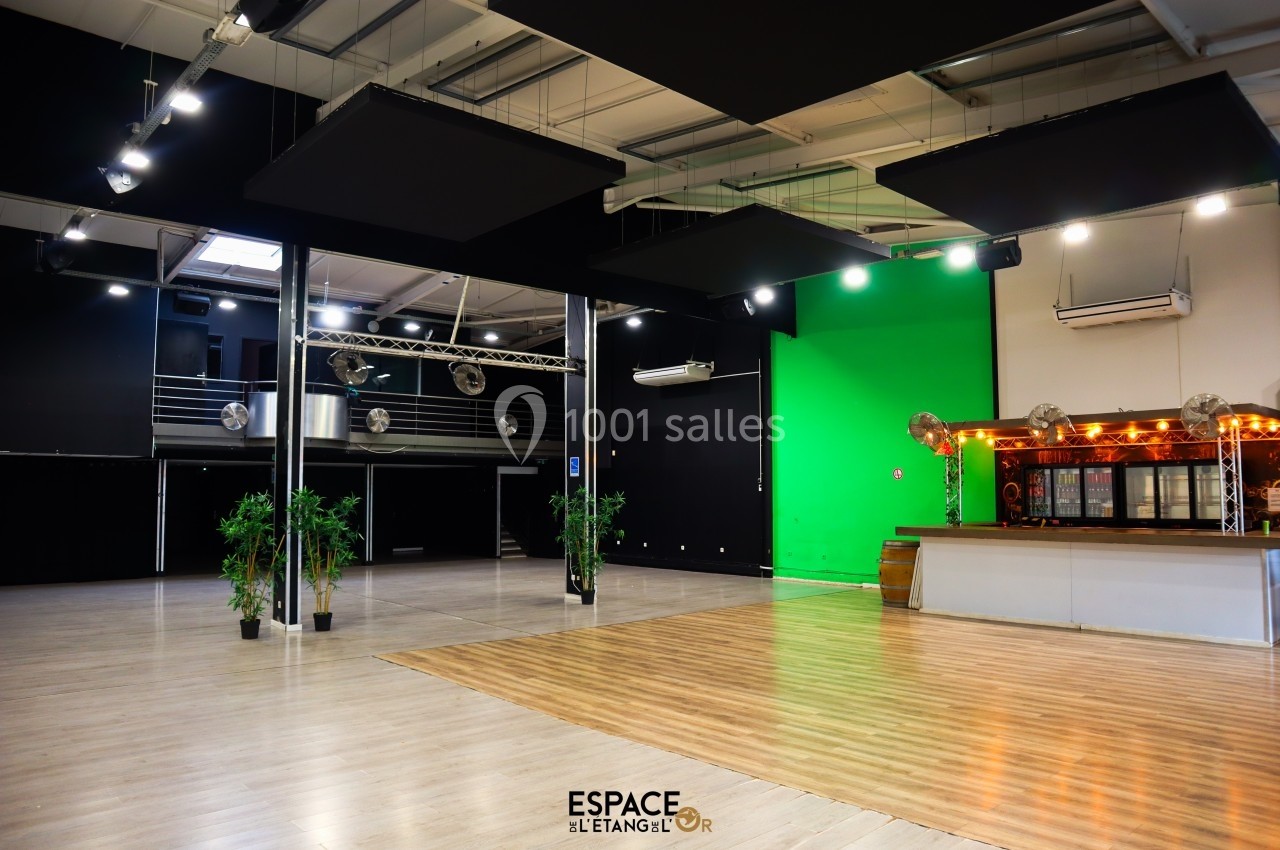 Salle spacieuse avec parquet, mur vert, bar éclairé et mezzanine, équipée pour événements ou tournages.