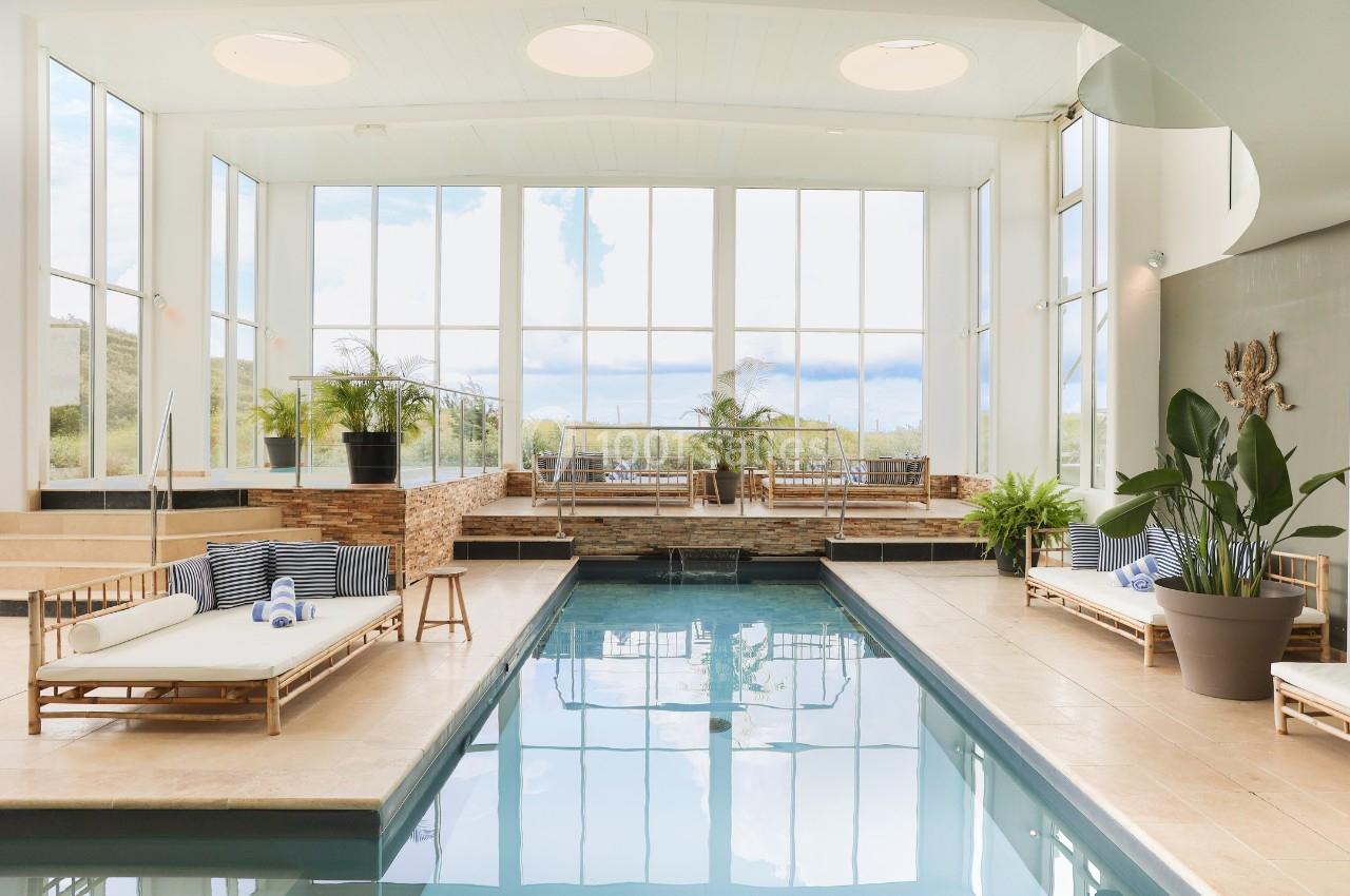 Piscine intérieure lumineuse avec grandes baies vitrées, entourée de chaises longues et de plantes décoratives.