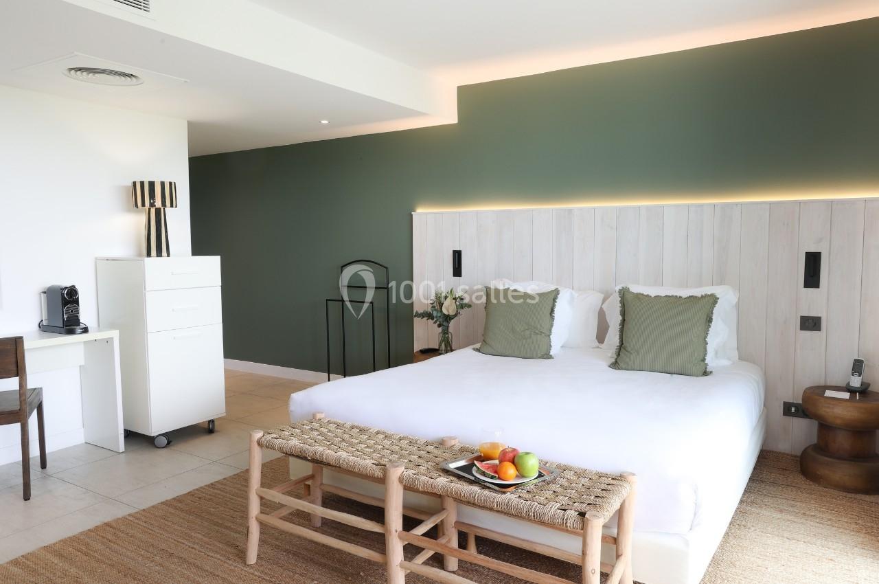 Chambre lumineuse avec lit double, mur vert, bureau, commode blanche, et panier de fruits sur un banc en osier.